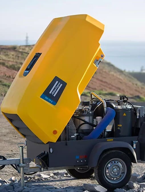 Compresor de aire portátil Atlas Copco con carcasa amarilla abierta, mostrando su motor interno. Montado sobre un remolque de dos ruedas, diseñado para aplicaciones en construcción y obras en exteriores.